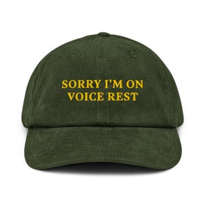 May include: A dark green corduroy baseball cap with the text "SORRY I'M ON VOICE REST" embroidered in yellow.