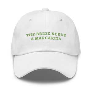 Puede incluir: Gorra de béisbol blanca con el texto bordado "THE BRIDE NEEDS A MARGARITA" en verde. La gorra tiene una visera curva y un botón en la parte superior. Un accesorio divertido para una despedida de soltera.