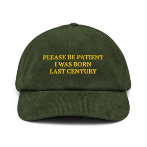 May include: Dark green corduroy baseball cap featuring the embroidered phrase "PLEASE BE PATIENT I WAS BORN LAST CENTURY" in gold. The cap has a curved peak and a structured crown.