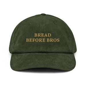 Puede incluir: Gorra de béisbol de pana verde oliva con la frase bordada "BREAD BEFORE BROS" en color dorado. La gorra tiene una visera curva y una corona estructurada. Un accesorio casual.