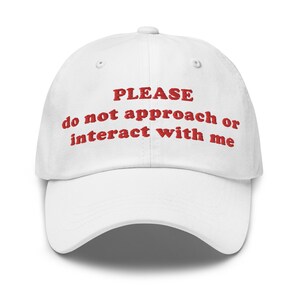 Op de afbeelding: Witte baseballcap met de rode geborduurde tekst "PLEASE do not approach or interact with me". De cap heeft een gebogen klep en een knoop aan de bovenkant.