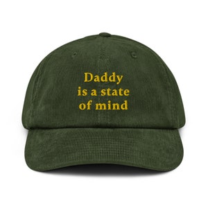 May include: A dark green corduroy baseball cap with the embroidered text "Daddy is a state of mind" in yellow. The cap has a curved brim and a structured crown, suitable for casual wear.