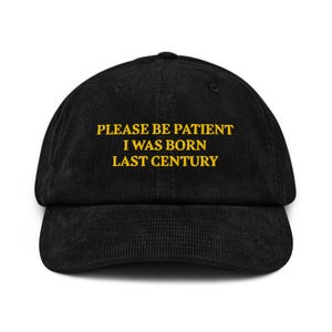 May include: Black corduroy baseball cap with a curved brim. The cap features the embroidered text "PLEASE BE PATIENT I WAS BORN LAST CENTURY" in gold thread. The hat has a classic six-panel design and an adjustable strap.