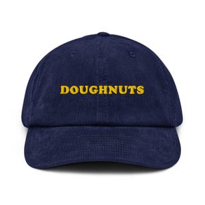 Puede incluir: Gorra de béisbol de pana azul marino con la palabra "DOUGHNUTS" bordada en amarillo en la parte delantera. La gorra tiene una visera curva y una corona estructurada.