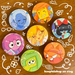 Op de afbeelding: Een verzameling van zes ronde buttons met kleurrijke illustraties in anime-stijl. De ontwerpen bevatten een blauwe figuur, een geel personage, een donkerharige figuur met een rode strik, een roze personage, een groene figuur en een oranje personage.