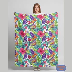 Pode incluir: Uma manta vibrante com um padrão tropical de beija-flores, flores de hibisco e folhagem verde exuberante. O design inclui tons de rosa, roxo, laranja e verde-azulado sobre um fundo branco. A manta é segurada por uma pessoa.