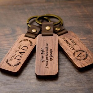 May include: Three wooden keychains with leather straps and metal rings. The keychains are engraved with the text "World's Best Dad", "Drive Safe! I need you here with me. Jennifer", and "Allison's Keys".