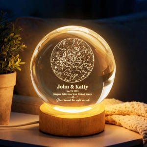 Op de afbeelding: Een heldere kristallen bol met een sterrenbeeldontwerp, verlicht vanaf een houten basis. De tekst "John & Katty, Jan 23, 2021, Niagara Falls, New York, United States, Stars danced the night we met" is gegraveerd.