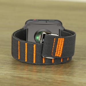 Op de afbeelding: Een zwarte smartwatch met een grijze elastische band met verticale oranje strepen. De band is vastgezet met een zilveren gesp. Het horloge ligt op een houten oppervlak.