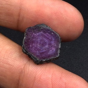 Könnte beinhalten: Ein roher, sechseckiger Edelstein mit einem tiefen violetten und magentafarbenen Farbton. Der Stein weist auf seiner flachen Oberfläche ein ausgeprägtes, natürlich vorkommendes konzentrisches geometrisches Muster mit dunklen, rauen Kanten auf.