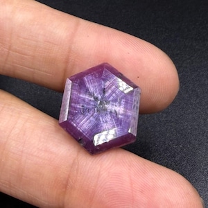 Puede incluir: Una piedra preciosa hexagonal y translúcida de color púrpura que presenta un patrón de estrella de seis puntas en su superficie facetada. La piedra muestra un tono violeta intenso con inclusiones internas visibles y acabado pulido.