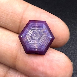 Könnte beinhalten: Ein sechseckig geschliffener violetter Edelstein mit einem ausgeprägten geometrischen Muster aus konzentrischen Linien, die von der Mitte ausstrahlen. Der Stein zeigt einen tiefvioletten Farbton mit sichtbaren inneren Streifen.