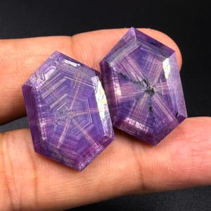 May include: Two faceted purple gemstones with a distinct hexagonal shape and a visible internal star-like pattern radiating from the center. The stones feature geometric lines and a polished surface that reflects light across their deep violet hues.