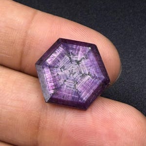 May include: A hexagonal-cut purple gemstone featuring a distinct six-rayed star pattern radiating from the center. The surface displays a translucent quality with visible internal linear inclusions and a polished, flat top facet.