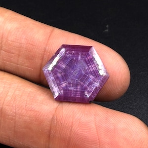 May include: A hexagonal-cut gemstone with a deep purple and magenta hue, featuring a distinct geometric pattern of concentric lines radiating from the centre. The stone has a polished, faceted surface that reflects light across its six sides.