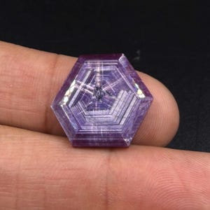 Puede incluir: Una piedra preciosa hexagonal de color púrpura con un patrón geométrico distintivo de líneas concéntricas que irradian desde el centro. La piedra muestra cualidades translúcidas con tonos violetas y magenta profundos y una apariencia estructurada.