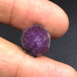 May include: A hexagonal, deep purple star ruby gemstone featuring a distinct six-rayed star pattern visible on its polished, flat surface. The stone displays a rich, saturated colour with natural inclusions and a dark, rough outer edge.