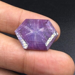 Può includere: Una gemma esagonale viola traslucida caratterizzata da un distinto motivo a stella a sei punte sulla superficie. La pietra presenta una parte superiore piatta e lucida con bordi sfaccettati e mostra una tonalità viola intenso.