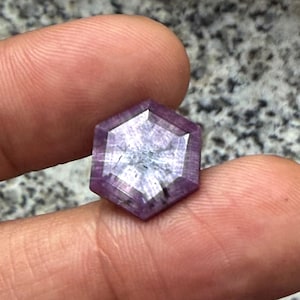 May include: A hexagonal cut gemstone with a deep purple and magenta hue, featuring a faceted surface that displays a star-like light reflection pattern across its center and visible internal inclusions within the translucent stone structure.