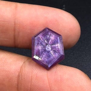 以下が含まれることがあります： ファセット加工された表面に6つの光条を持つ星模様が特徴の六角形の紫色の宝石。この石は深いバイオレットの色合いを見せ、交差する光の線が半透明の結晶構造の中に幾何学的な星の効果を生み出しています。