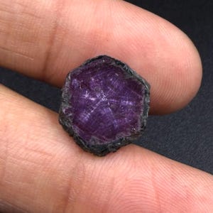 Könnte beinhalten: Ein roher, sechseckiger Edelstein mit einem tiefen violetten und magentafarbenen Farbton. Die Oberfläche des Steins zeigt ein ausgeprägtes, natürliches sternförmiges Muster, das von der Mitte ausstrahlt, mit einem dunklen, rauen und texturierten Rand.
