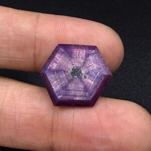Puede incluir: Una piedra preciosa de corte hexagonal con un tono púrpura y magenta intenso, que presenta un patrón de estrella de seis rayos que irradia desde una inclusión central oscura. La piedra es translúcida con líneas de crecimiento internas visibles.