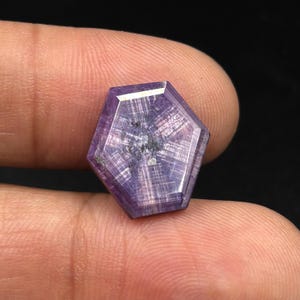 Puede incluir: Una piedra preciosa hexagonal de tono púrpura con un patrón distintivo en forma de estrella que irradia desde el centro. La piedra presenta una superficie facetada con inclusiones internas visibles y un acabado pulido de forma geométrica.