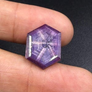 Könnte beinhalten: Ein hexagonaler violetter Edelstein mit einem ausgeprägten sechsstrahligen Sternmuster auf der Oberfläche. Der Stein zeigt tiefe violette Farbtöne mit helleren, sich kreuzenden Linien, die einen geometrischen Sterneffekt erzeugen.