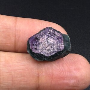 Könnte beinhalten: Ein roher, sechseckiger Edelstein mit einem tiefvioletten Zentrum, das zu dunklen, undurchsichtigen Rändern übergeht. Die Oberfläche zeigt ein ausgeprägtes geometrisches, gitterartiges Kristallmuster mit sichtbaren Streifen und einer natürlichen Textur.
