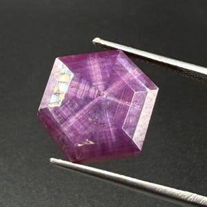 May include: A hexagonal, faceted purple gemstone with a distinct star-like pattern radiating from the centre. The stone displays translucent qualities and internal linear inclusions, held by silver-toned metal tweezers against a dark background.