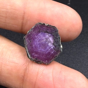 Op de afbeelding: Een ruwe, zeshoekige robijn met een diepe magenta en paarse tint. De steen heeft een natuurlijk, ongepolijst oppervlak met zichtbare kristallijne strepen en een donkere, ruwe rand, wat de authentieke, onbehandelde minerale structuur laat zien.