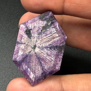 Può includere: Una gemma sfaccettata di forma esagonale che mostra una tavolozza di colori viola e magenta. La pietra presenta un motivo a stella che si irradia dal centro, con inclusioni scure visibili e trame lineari che si intersecano.