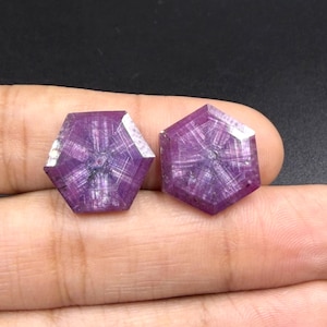 Op de afbeelding: Twee zeshoekig geslepen robijnen met een diepe magenta en paarse tint. Elke edelsteen vertoont een duidelijk zonnestraalpatroon met zes stralen vanuit het midden, met zichtbare interne insluitingen en facetten die het licht vangen.