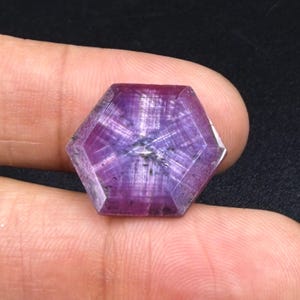 Può includere: Una gemma esagonale traslucida di colore viola con un distinto motivo a stella a sei punte sulla superficie sfaccettata. La pietra mostra intense tonalità magenta e viola con inclusioni interne visibili e un taglio geometrico.