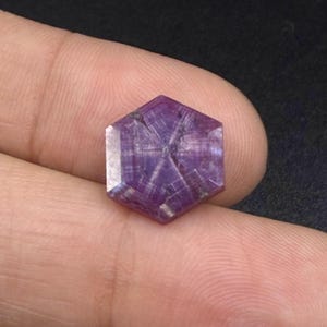 May include: A hexagonal, translucent purple gemstone featuring a distinct six-rayed star pattern radiating from the center. The stone displays a polished, flat surface with visible internal fibrous inclusions that create the star effect.