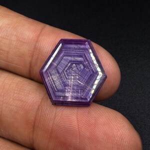 以下が含まれることがあります： 明確な同心円状の幾何学模様と線状のインクルージョンが特徴の六角形の紫色の宝石。深い紫の色合いを持つファセットカットされた表面と磨き上げられた仕上げが施されており、結晶内部の対称的な構造が際立っています。