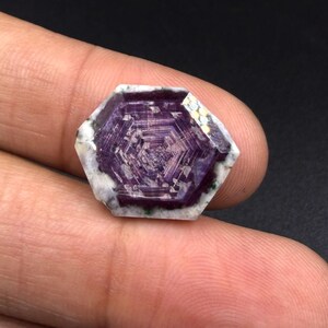 May include: A hexagonal-cut gemstone featuring a deep purple centre with a distinct geometric star pattern, surrounded by a white and grey mottled border. The stone displays a polished, flat surface with visible internal crystalline structures.