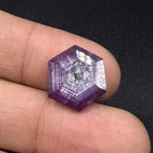 Könnte beinhalten: Ein hexagonaler violetter Edelstein mit ausgeprägten konzentrischen geometrischen Mustern und einer facettierten Oberfläche. Der Stein zeigt tiefe violette Farbtöne mit helleren, durchscheinenden Abschnitten und einem dunklen, strukturierten Kern.