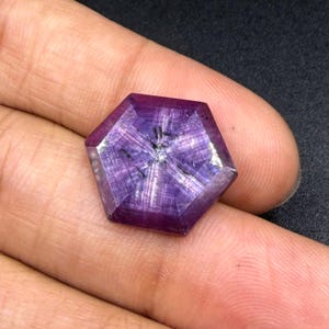May include: A hexagonal purple gemstone featuring a distinct six-rayed star pattern known as asterism. The stone displays a translucent, deep violet hue with visible internal fibrous inclusions that create a shimmering, light-reflecting effect.