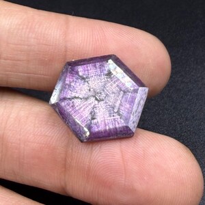 Könnte beinhalten: Ein hexagonaler violetter Edelstein mit einem ausgeprägten sechsstrahligen Sternmuster auf der Oberfläche. Der Stein zeigt einen tiefvioletten Farbton mit sichtbaren inneren Streifen und einer polierten Oberfläche.