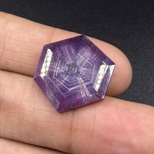 May include: A hexagonal purple gemstone featuring a distinct geometric pattern of concentric lines radiating from the center. The surface displays a polished finish with two elongated, light-reflecting facets on opposite sides of the stone.