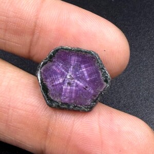 Könnte beinhalten: Ein sechseckiger violetter Edelstein mit einem ausgeprägten sternförmigen Muster, das von der Mitte ausstrahlt. Der Stein hat einen dunklen, strukturierten und unebenen Rand, der die leuchtend violette Fläche umgibt und natürliche Einschlüsse zeigt.