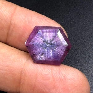 Könnte beinhalten: Ein sechseckiger, facettierter Edelstein in einem tiefen Violett- und Magentaton. Der Stein zeigt ein ausgeprägtes sternförmiges Muster, das von der Mitte ausstrahlt, mit sichtbaren inneren Einschlüssen und einer polierten, reflektierenden Oberfläche.