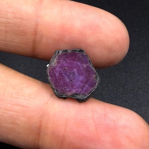 May include: A raw, hexagonal-shaped gemstone with a deep magenta and purple hue. The stone features a distinct six-sided geometric pattern on its flat surface, with dark, rough edges surrounding the vibrant, crystalline center of the mineral.