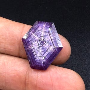 Könnte beinhalten: Ein facettierter violetter Edelstein mit einem ausgeprägten hexagonalen internen Wachstumsmuster. Der Stein hat eine polierte Oberfläche mit sichtbaren geometrischen Linien, die von der Mitte ausstrahlen und tiefe violette Farbtöne zeigen.
