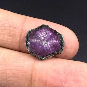 Può includere: Una fetta di pietra preziosa grezza e non lucidata con un centro magenta e viola intenso che presenta un distinto motivo a stella. La pietra ha un bordo esterno ruvido, scuro e irregolare, che mostra una texture cristallina naturale.