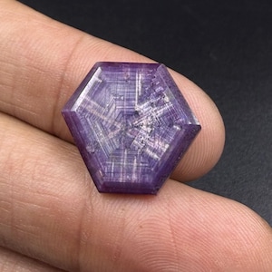 Può includere: Una gemma esagonale di colore viola caratterizzata da un distinto motivo a stella a sei punte sulla superficie. La pietra è traslucida con visibili inclusioni lineari interne e una superficie superiore piatta e lucida con bordi netti.