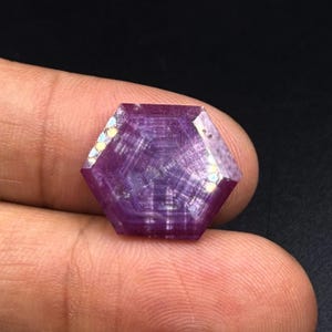 Könnte beinhalten: Ein sechseckig geschliffener Edelstein in tiefen Violett- und Magentatönen mit einem ausgeprägten geometrischen Innenmuster aus konzentrischen Linien und einer facettierten Oberfläche, die das Licht auf allen sechs Seiten reflektiert.
