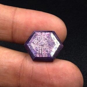 Könnte beinhalten: Ein sechseckig geschliffener violetter Edelstein mit einem ausgeprägten geometrischen Muster aus konzentrischen Linien, die von der Mitte ausstrahlen. Der Stein weist eine durchscheinende Qualität mit sichtbaren inneren Einschlüssen und eine polierte, facettierte Oberfläche auf.