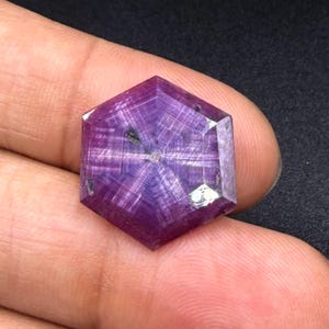 May include: A hexagonal, faceted gemstone with a deep purple and magenta hue, displaying a distinct six-rayed star pattern radiating from the center. The stone features visible internal inclusions and a polished surface with geometric facets.